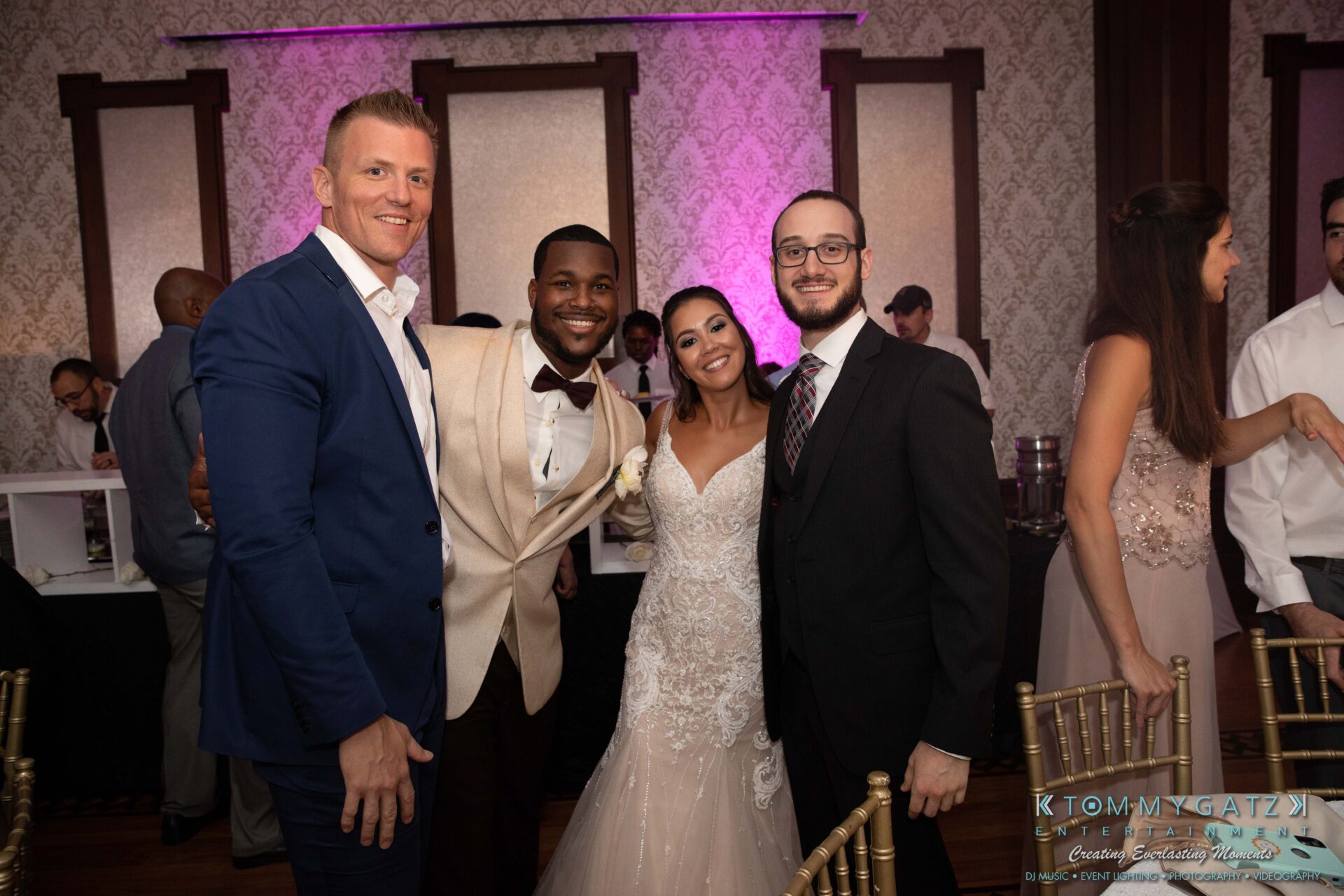 Four people dressed in formal wear, including a bride in a white gown and a groom in a cream suit, stand smiling together in a warmly lit room with patterned walls.