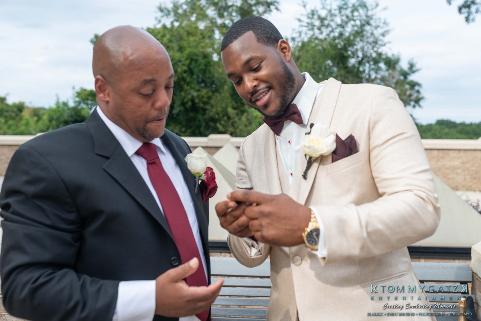 Two men in suits stand outdoors looking at a smartphone. One wears a black suit with a red tie, the other a beige suit with a burgundy tie and boutonnière.