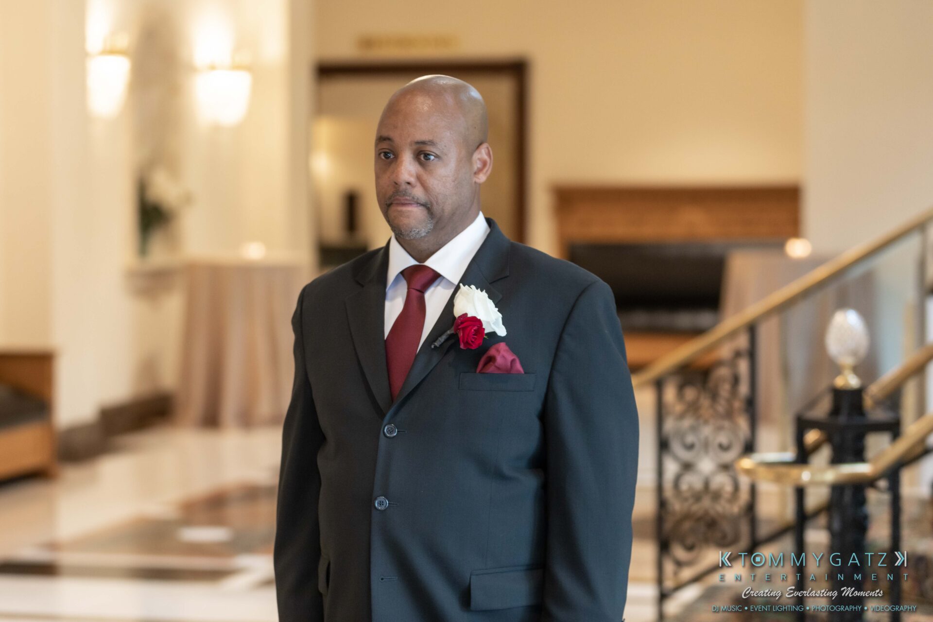 A man in a dark suit with a white shirt and red tie stands in a hotel lobby, wearing a boutonniere.