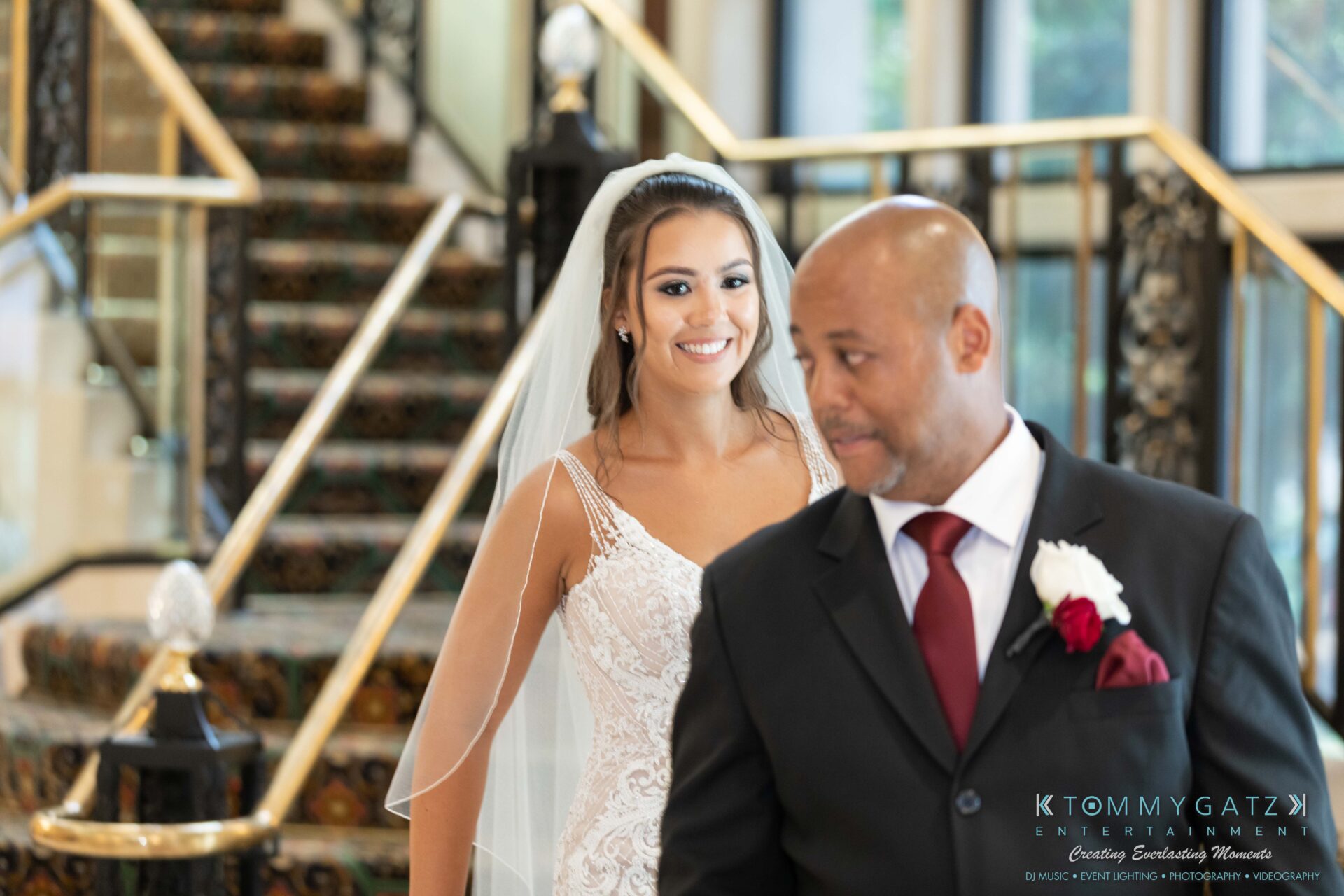 Bride in a white dress smiles while standing behind a man in a suit with a red tie and boutonnière, set against a staircase background.