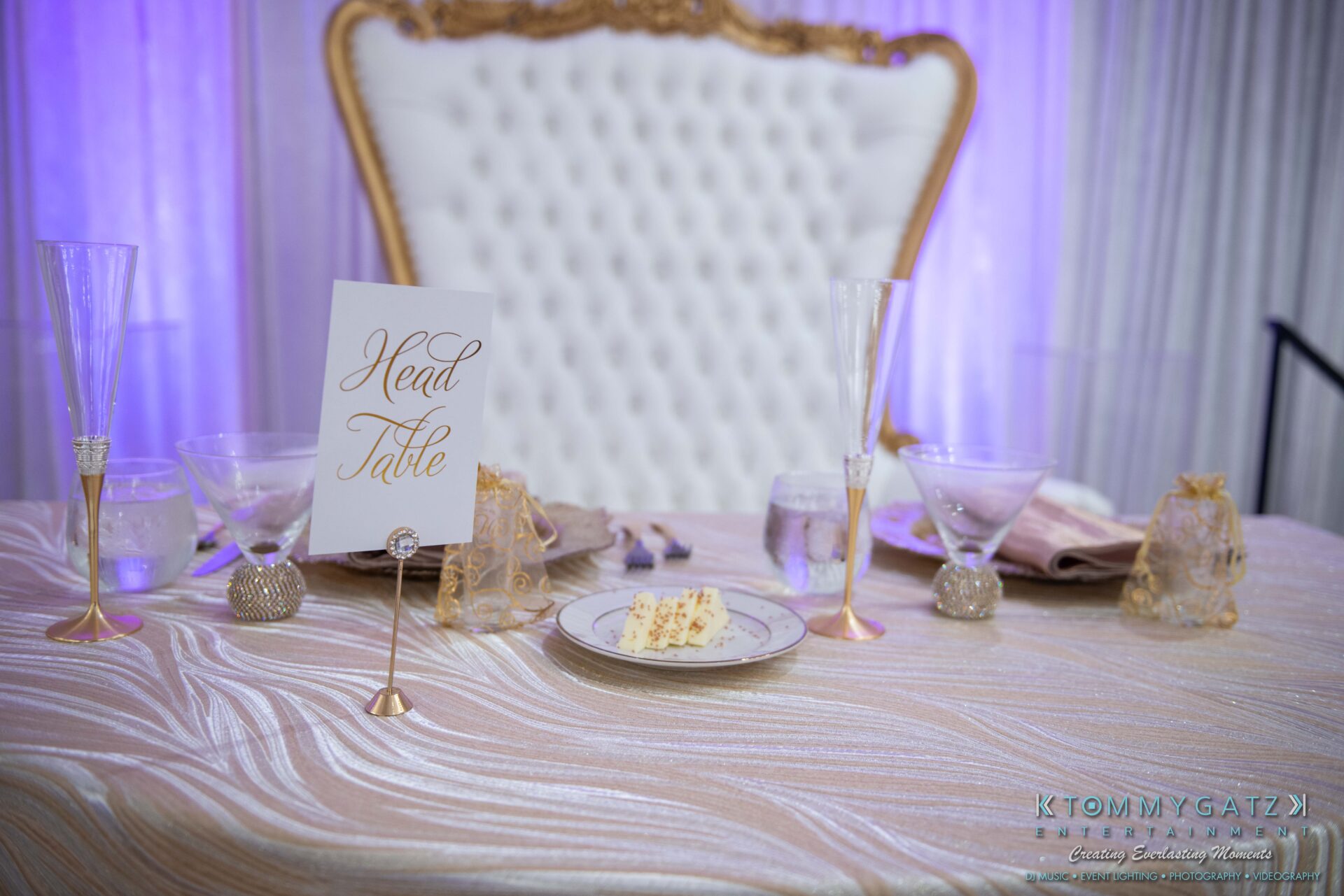 Elegant head table setup with a gold-accented chair, champagne glasses, a plate with cake slices, and a sign reading "Head Table" on a shimmering tablecloth.