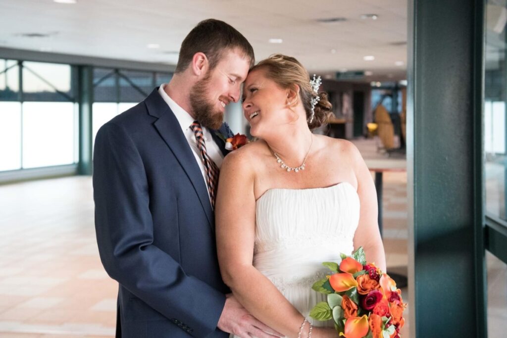A bride and groom share a moment indoors, smiling and leaning towards each other. The bride holds a bouquet of orange and red flowers.