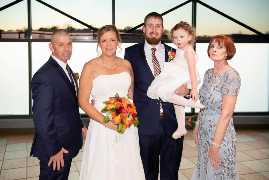 A wedding portrait with five people. A bride in a white dress holds a bouquet, standing beside a groom in a suit who is holding a child. Two older adults stand on either side, all posed indoors.