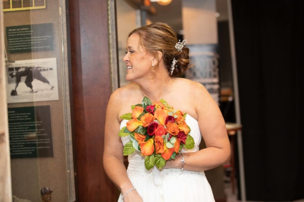 A bride holding a bouquet of orange and red flowers looks to her right, standing indoors next to a wall with framed photos and text.