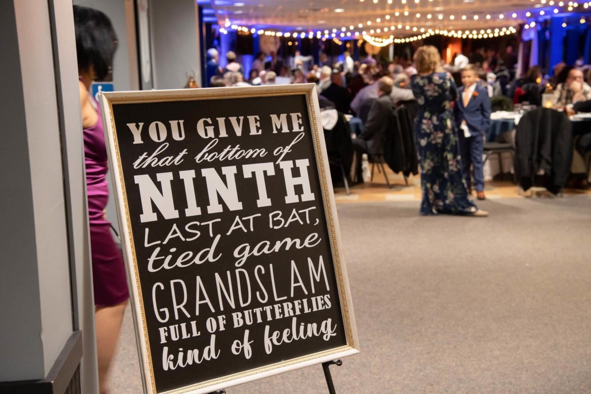 Sign with text about a baseball game feeling in the foreground; people socializing in a decorated venue in the background.