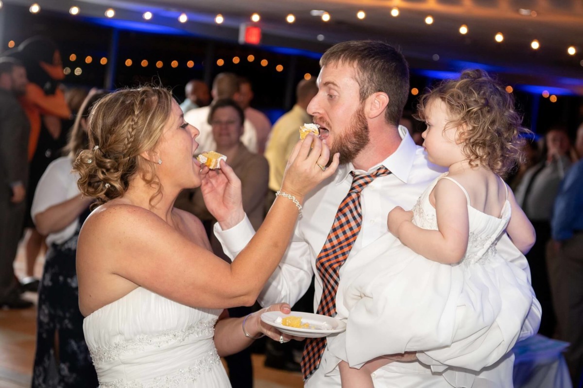 A bride and groom feed each other cake at a wedding reception. The groom holds a young child who looks on. Guests are visible in the background.