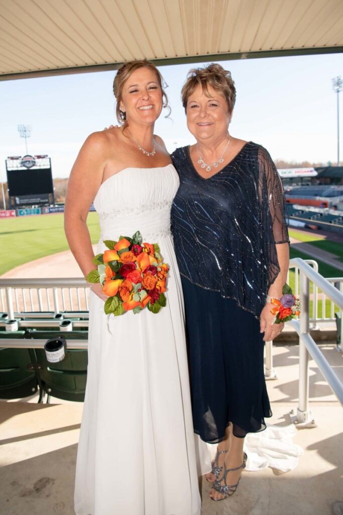 Two women smiling; one in a white dress holding a bouquet, the other in a dark dress. They're standing in a stadium with empty seats and a baseball field in the background.