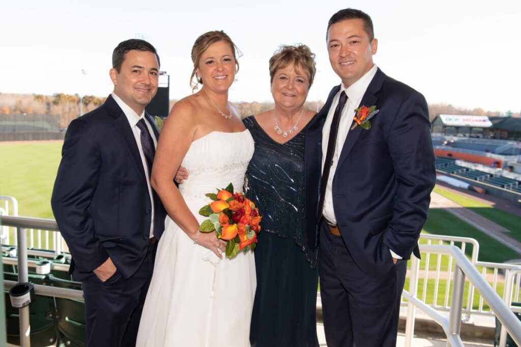 A bride in a white dress stands with two men in suits and a woman in a dark dress, holding a bouquet, with a sports field in the background.