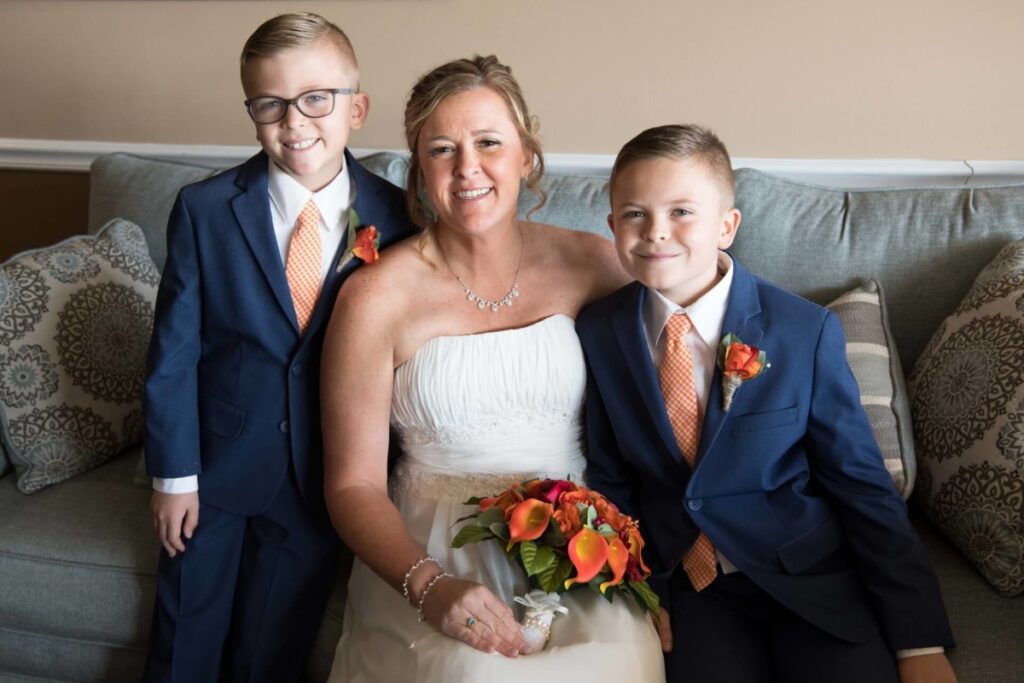 A smiling woman in a white dress sits on a couch between two young boys in blue suits with orange ties and boutonnieres.