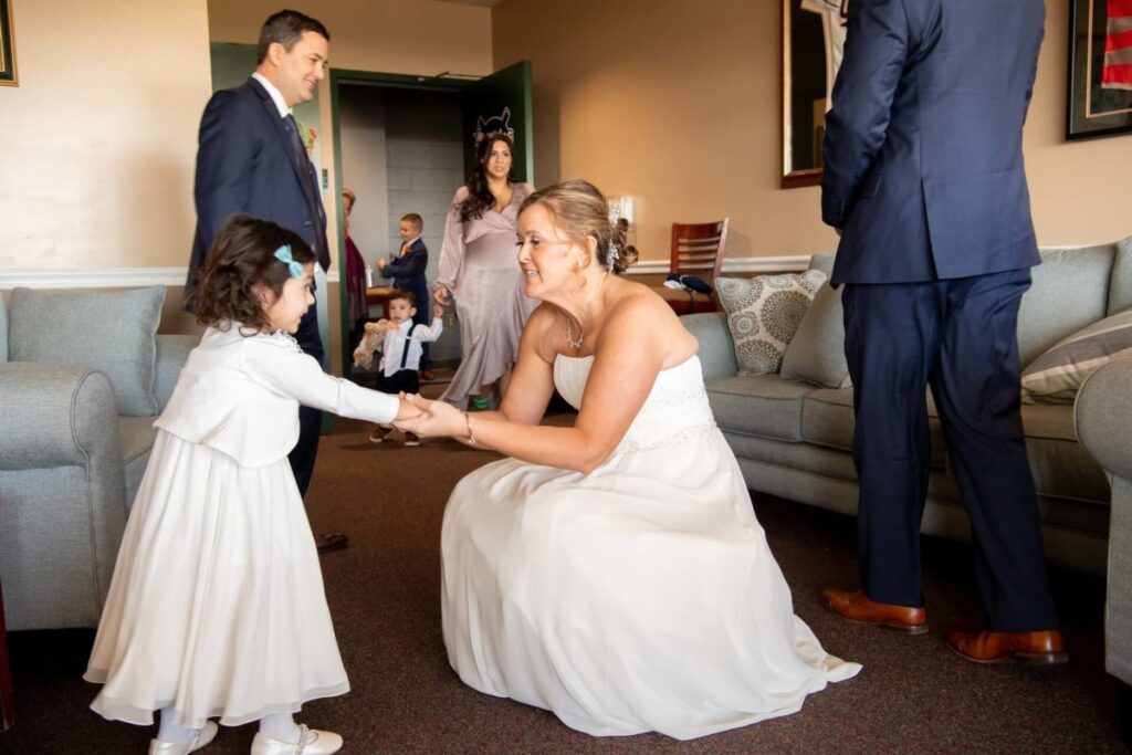 A bride in a white dress kneels to greet a young girl in a white outfit. Several adults and children are in the room, with some standing and some seated.