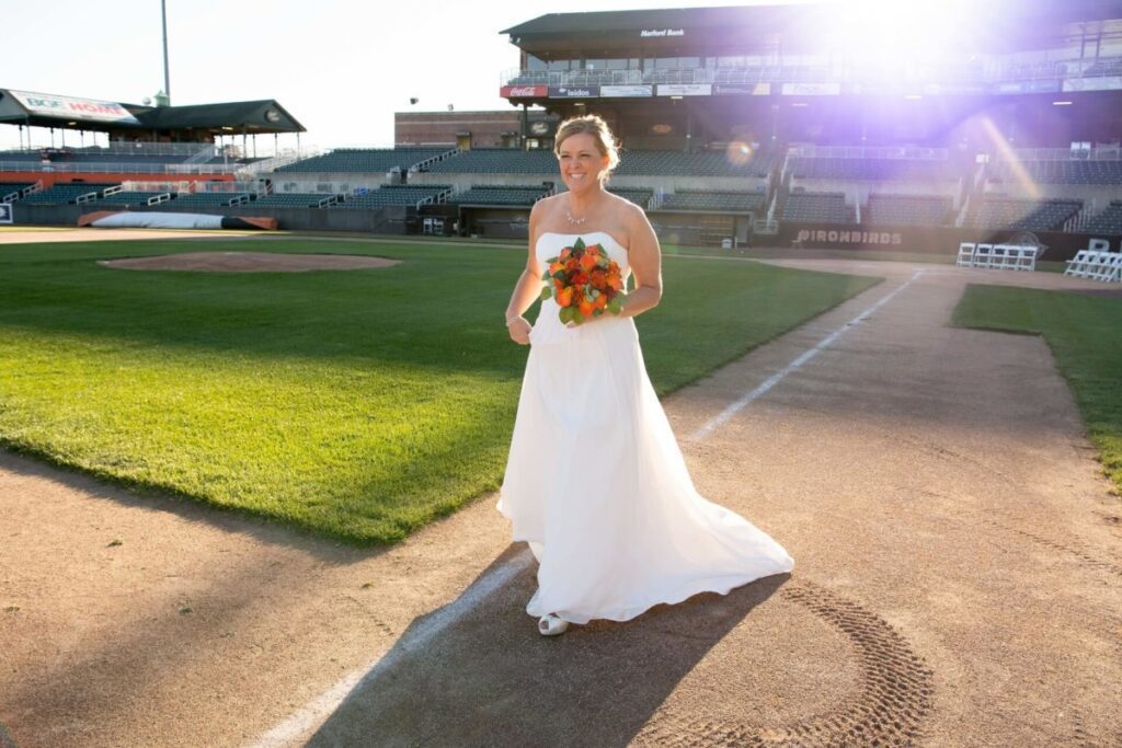 A bride in a white wedding dress walks on a baseball field, holding a bouquet of orange flowers. The sun is shining brightly in the background.