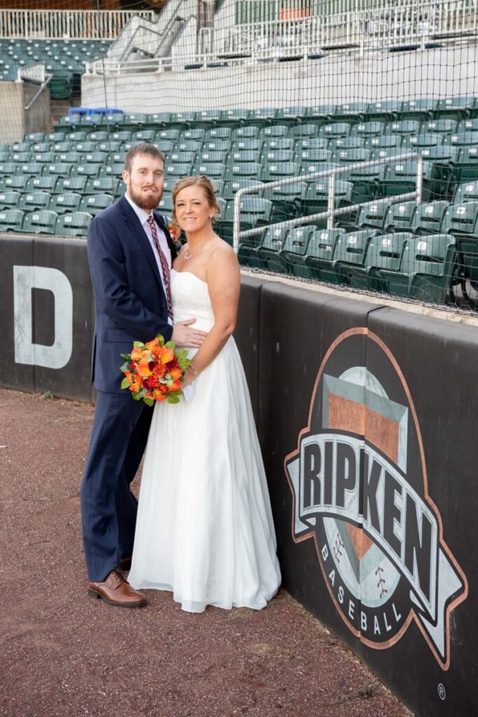 A bride and groom stand by a "Ripken Baseball" sign in a baseball stadium. The bride holds a bouquet.