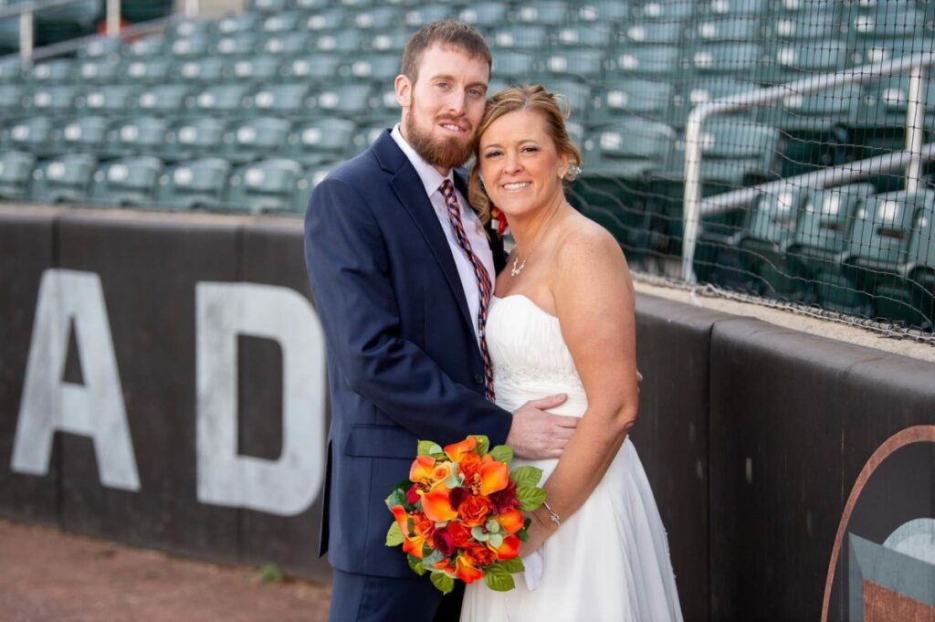 A couple in wedding attire stands together on a baseball field, with the bride holding a bouquet of orange flowers.