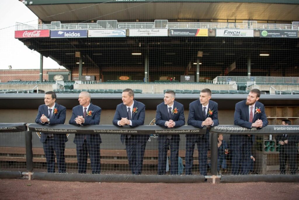 Six men in suits stand behind a baseball dugout fence, wearing boutonnieres.