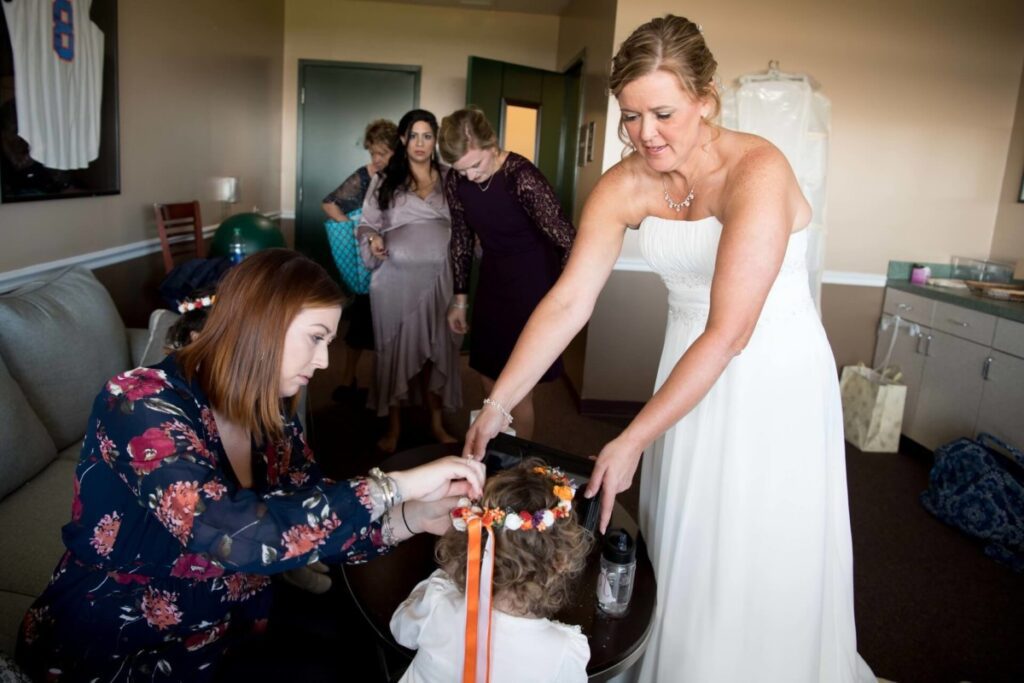 A bride in a white dress adjusts a child's hair with help from another woman in a floral blouse. Several people are in the background in a room.