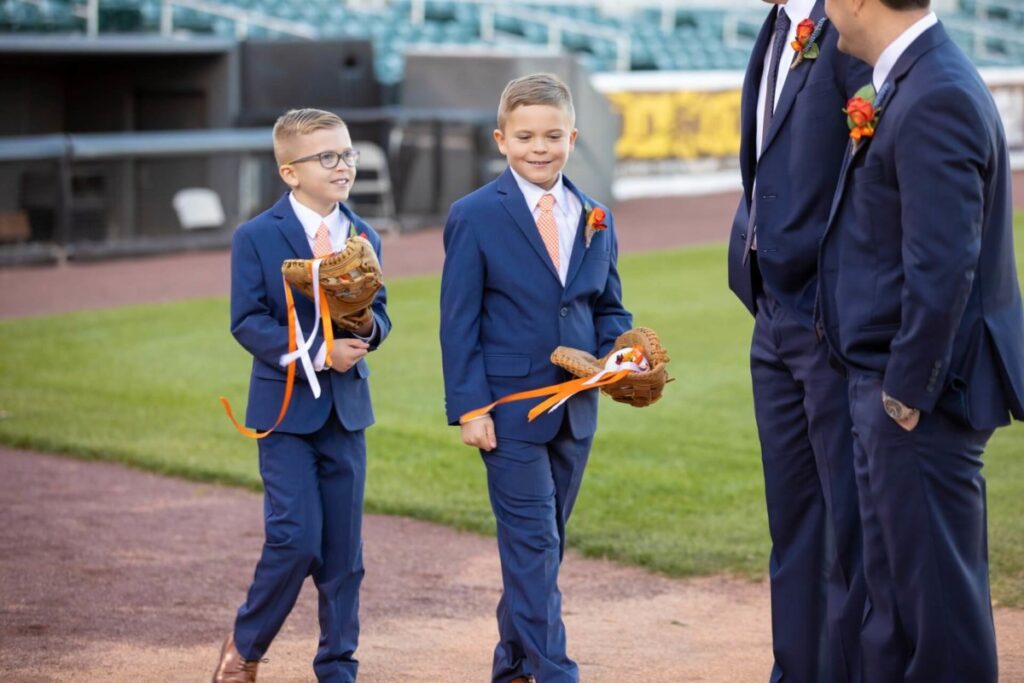 Two boys in blue suits walk on a baseball field holding gloves with orange ribbons. Two adults in similar attire walk beside them.