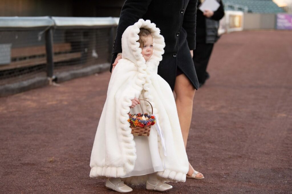 A child in a white, hooded cloak holds a basket of flowers. They stand on a path next to an adult in a dark coat.