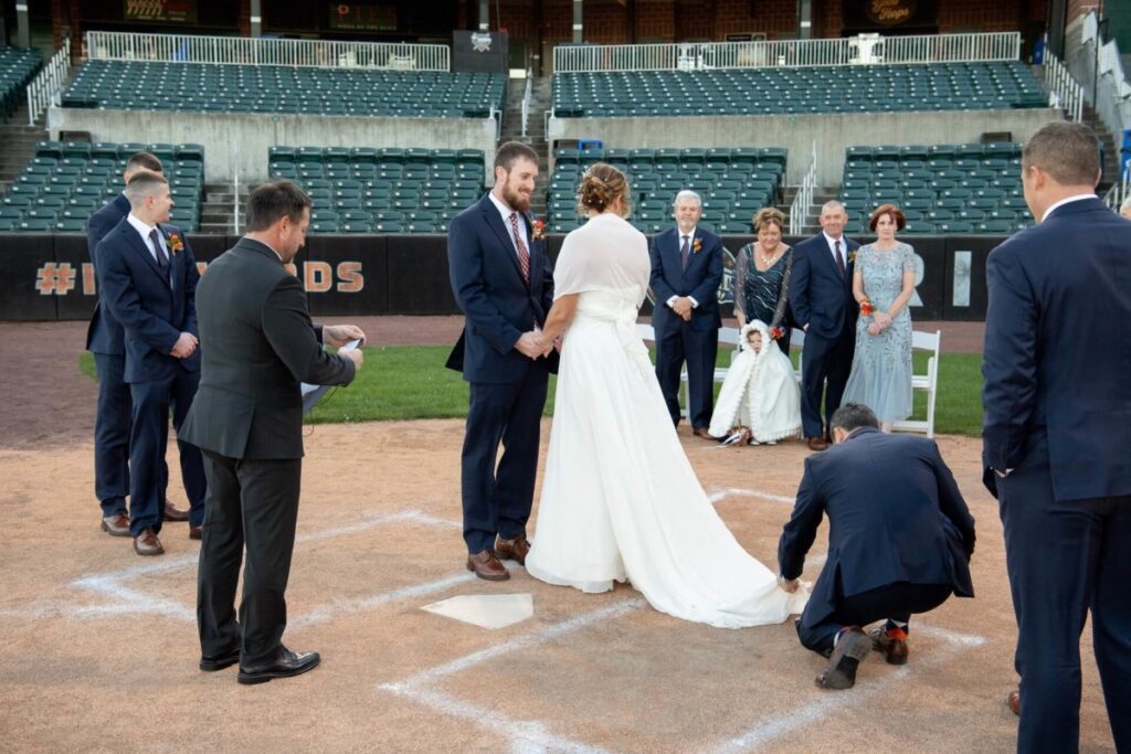 A couple exchanges vows in a baseball stadium, surrounded by their wedding party and family. One person kneels to adjust the bride's dress.