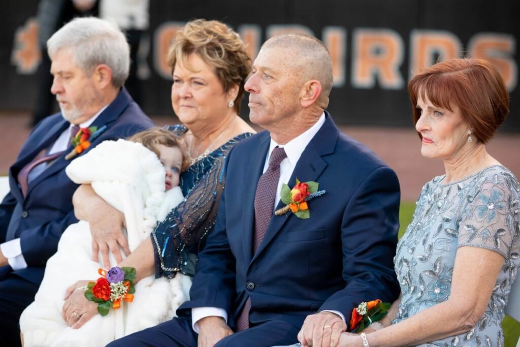 Four adults seated, dressed formally, with corsages. One holds a bundled child. They're outdoors with a blurred "BIRDS" sign in the background.