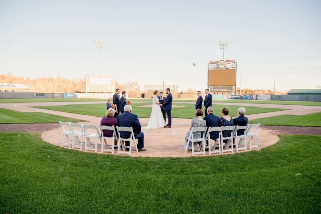 A wedding ceremony is taking place on a baseball field, with the couple standing on the pitcher's mound, surrounded by a small gathering of seated guests.