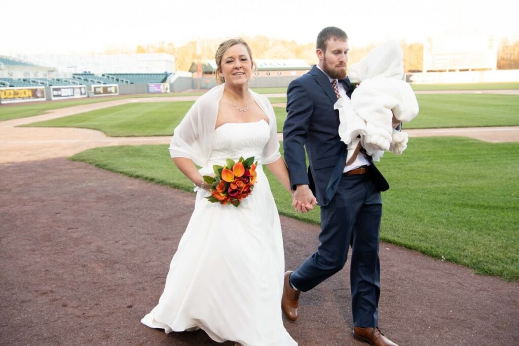 Bride and groom walking on a baseball field, holding hands. The bride holds a bouquet, and the groom carries a baby swaddled in a blanket.