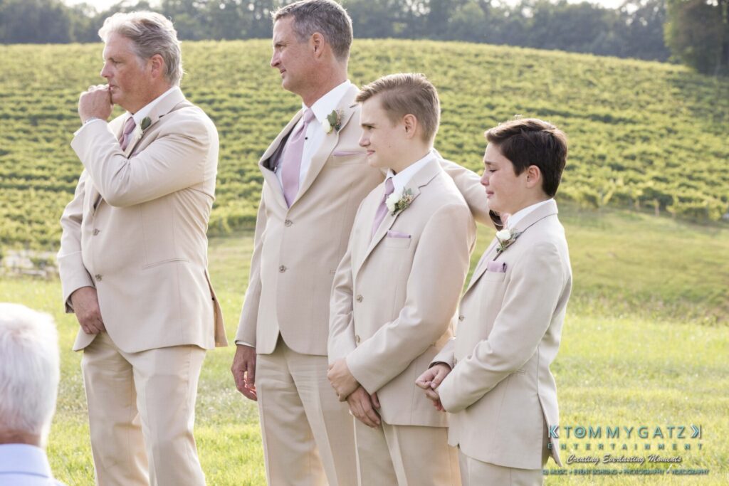 Four men in beige suits stand outdoors on grass with a vineyard in the background.