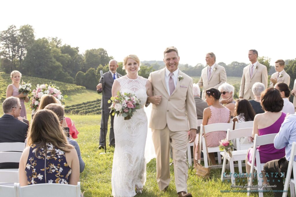 A couple walks down the aisle outdoors after a wedding ceremony, surrounded by guests seated on white chairs.