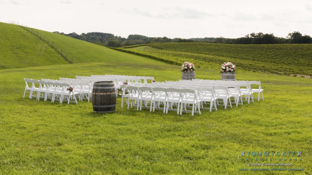 Outdoor wedding setup with rows of white chairs facing a barrel and floral arrangements, on a grassy hillside.