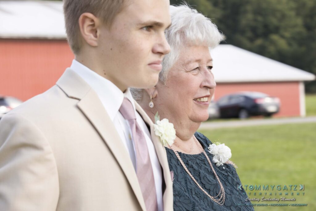 A young man in a beige suit and an older woman in a teal dress, both wearing floral boutonnieres, walk outdoors near a red building.
