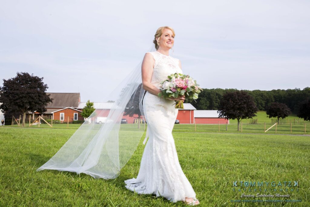 Bride in a white dress holding a bouquet, walking on a grassy field with barns and trees in the background.
