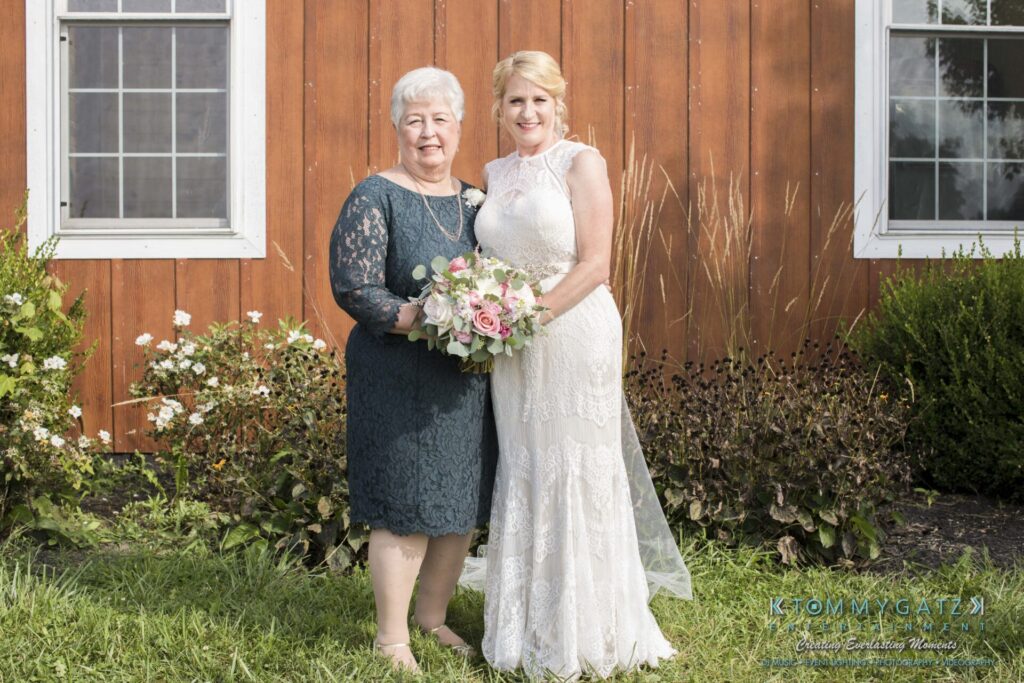 Two women stand side by side in front of a wooden building, both smiling. One woman holds a bouquet of flowers.