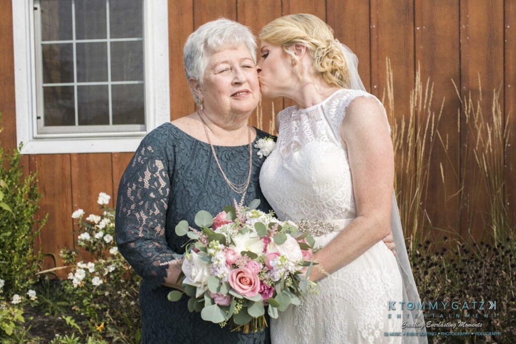 A woman in a wedding dress kisses an older woman on the cheek. The older woman holds a bouquet of flowers. They stand in front of a wooden wall with a window.