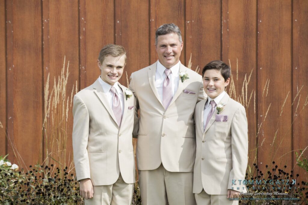 Three people in beige suits with lavender ties stand in front of a wooden wall, smiling.