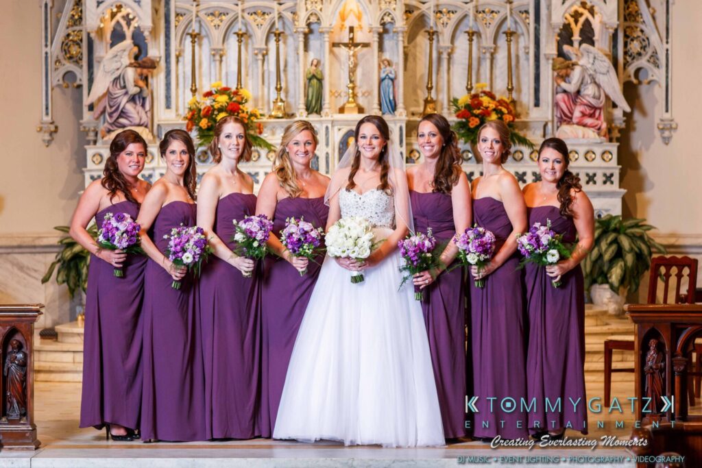 Bride in white with bridesmaids in purple dresses holding bouquets, standing in a decorated church.