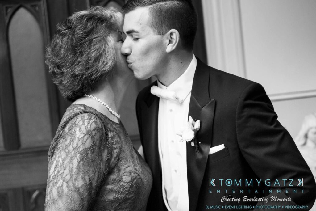 A groom kisses a woman on the cheek, both in formal attire, against a backdrop of wood and decorative paneling.