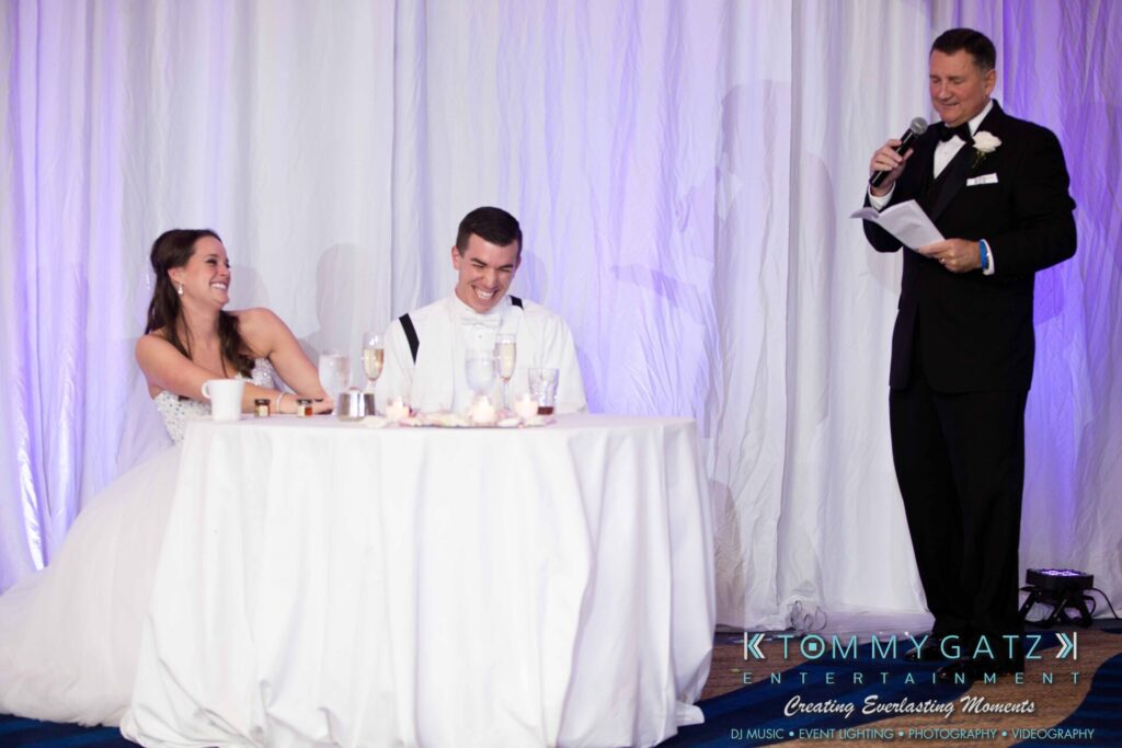 A bride and groom sitting at a table laugh as a man in a tuxedo speaks into a microphone.