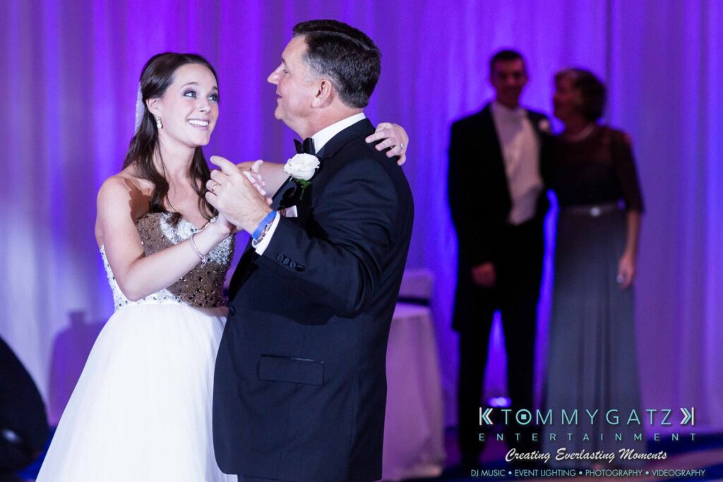 A bride and an older man share a dance at an event, with people observing in the background.
