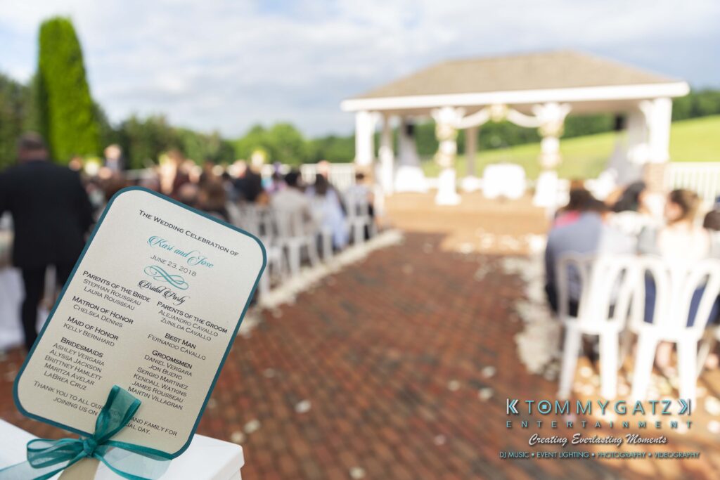 Outdoor wedding ceremony with chairs arranged on a brick path facing a decorated gazebo. A wedding program is in the foreground.
