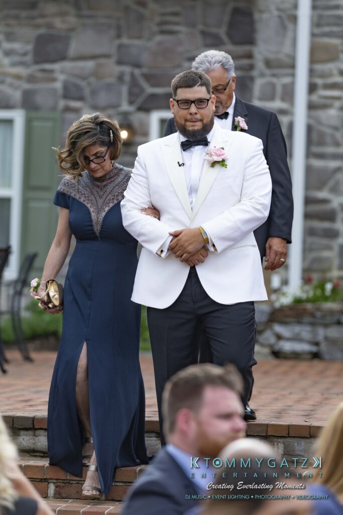 A man in a white tuxedo escorts a woman in a navy dress down brick steps. A man in a suit follows behind.