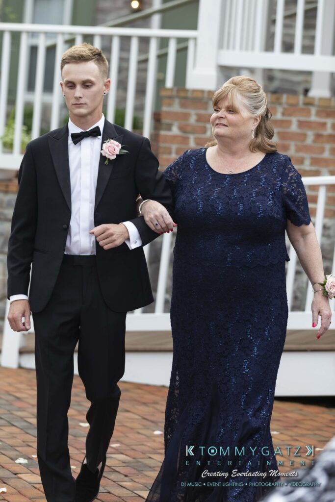 A young man in a suit escorts an older woman in a blue dress. They are walking outdoors on a brick path with a white railing behind them.