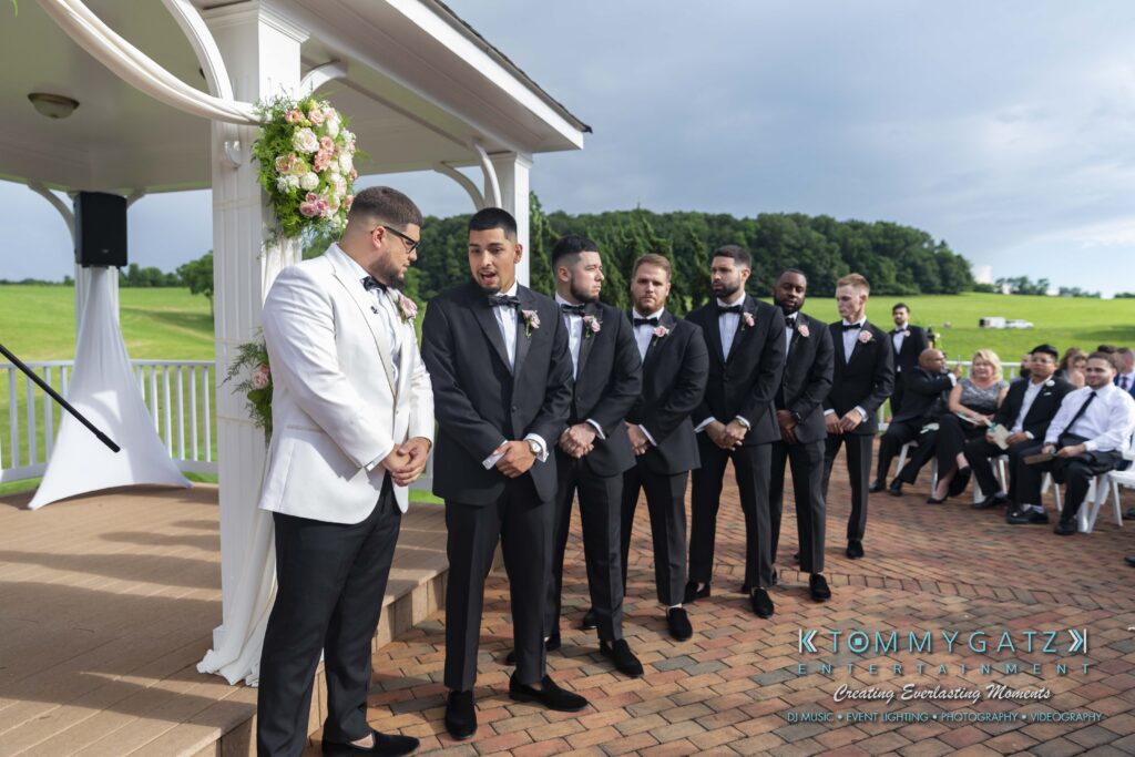 A groom in a white tuxedo and six groomsmen in black tuxedos stand under a floral-decorated gazebo on a sunny day, with guests seated in the background.