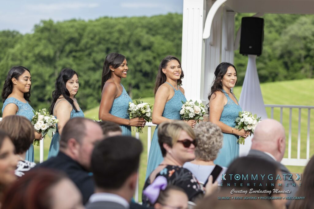 Bridesmaids in teal dresses stand holding bouquets at an outdoor wedding ceremony, with onlookers seated in the foreground.