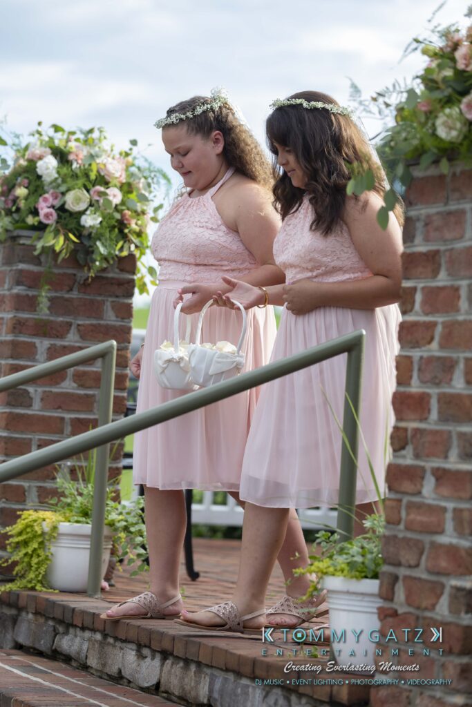 Two women in pink dresses walk down brick steps, holding a basket of white flowers. They're outdoors, with greenery and floral arrangements in the background.