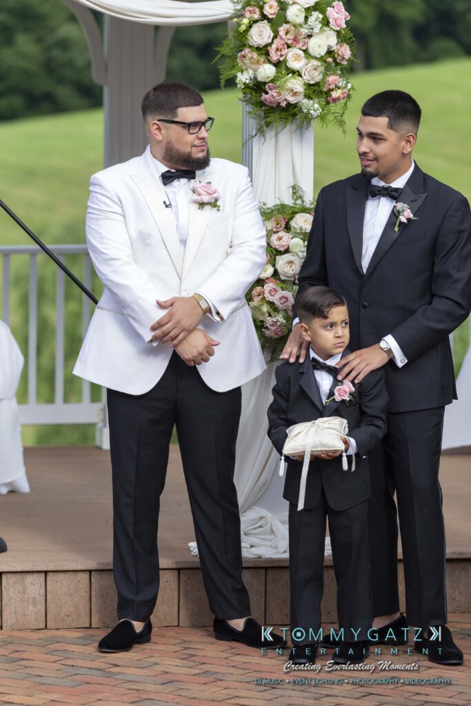 Three people in formal attire at an outdoor event; two men in suits with a young boy holding a ring pillow. Floral decorations in the background.