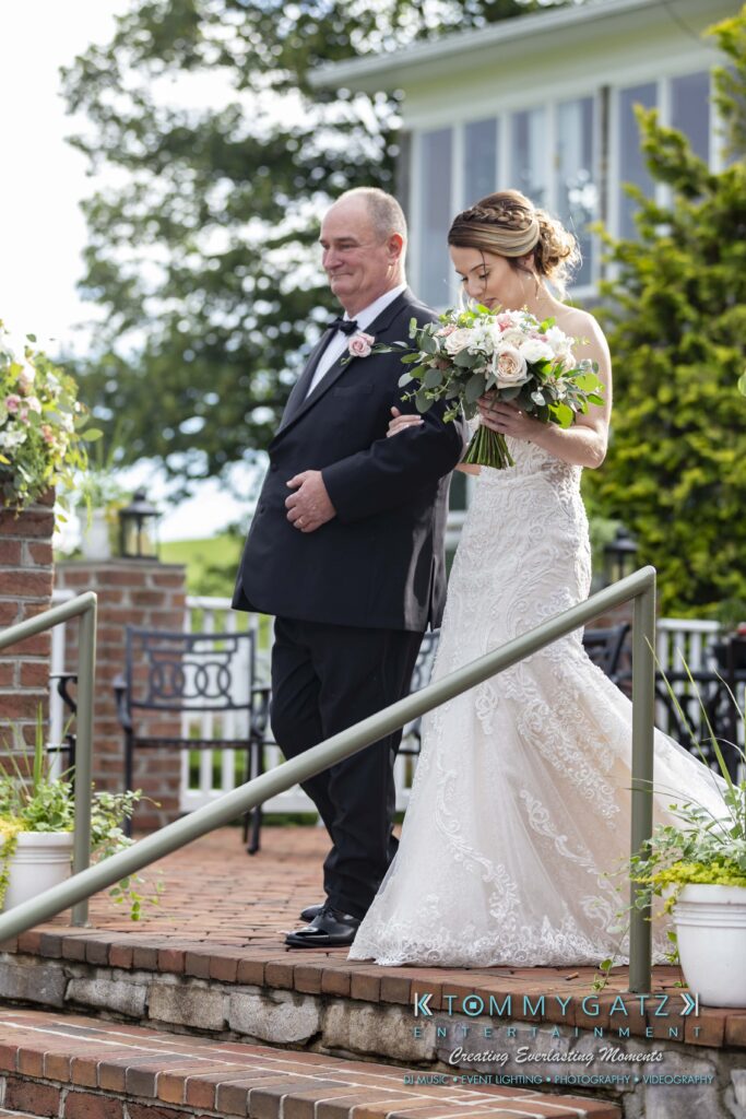 A bride in a strapless gown walks down steps with an older man in a black suit. She holds a bouquet of flowers. They are outside near a brick structure.