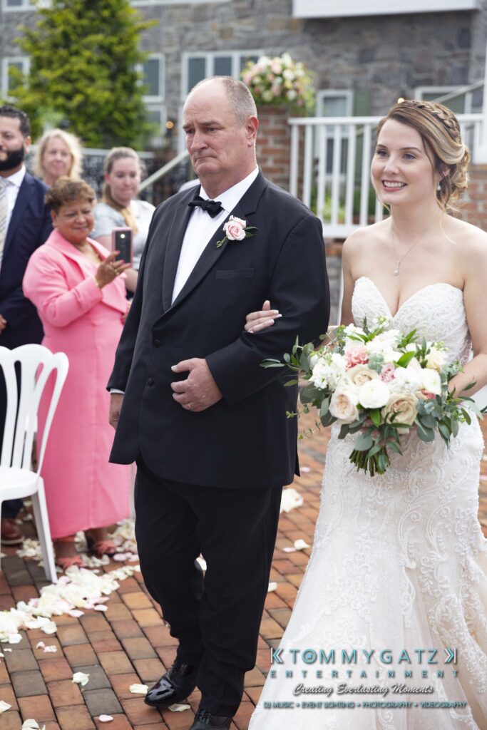 A bride in a white gown walks down an outdoor aisle with an older man in a black suit. She holds a bouquet and smiles. Guests and flower petals are visible.
