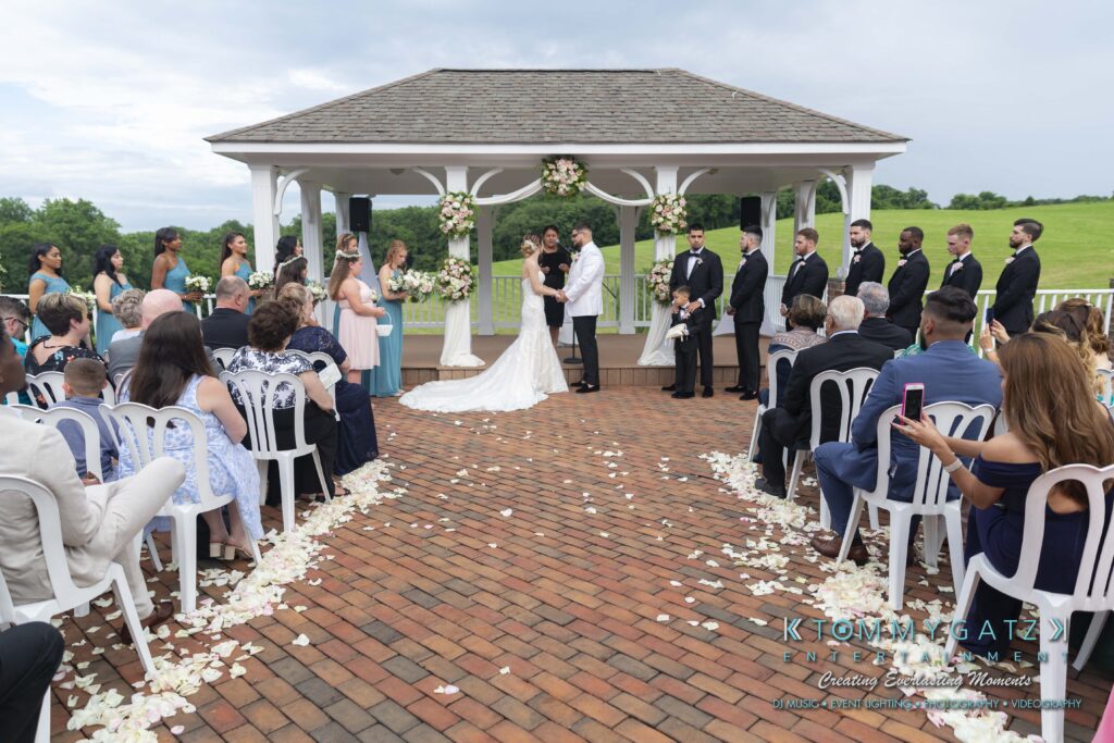 Outdoor wedding ceremony under a gazebo with a couple kissing. Guests are seated, and the wedding party stands on either side. White chairs and flower petals line the brick path.