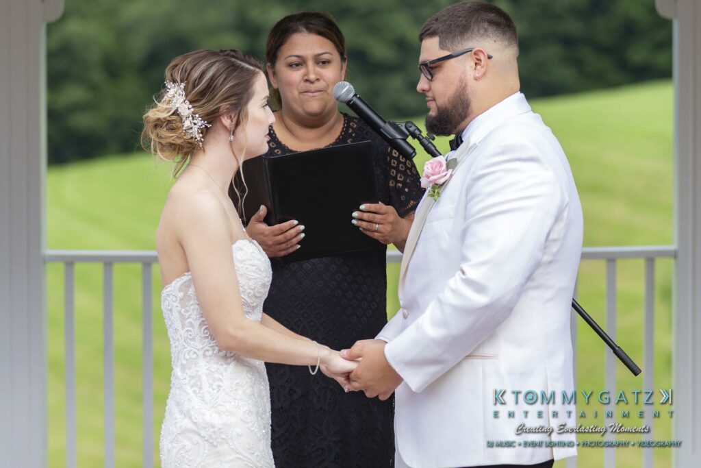 Couple holding hands during a wedding ceremony under an outdoor gazebo, with an officiant speaking behind a microphone.