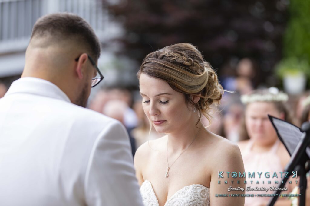 A couple in formal attire stands together at an outdoor ceremony. The woman has braided hair and closed eyes. The man is wearing a white suit. People are visible in the background.