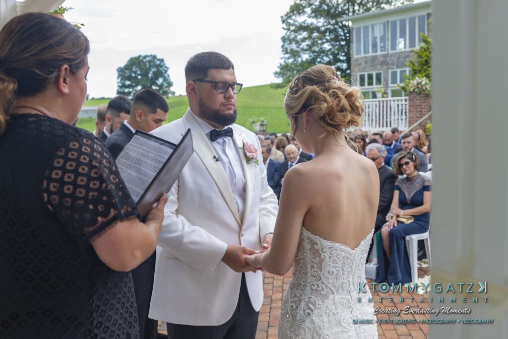 A couple holds hands during an outdoor wedding ceremony, with an officiant reading from a folder. Guests are seated in the background.